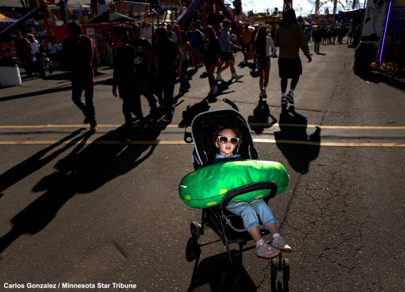 A young child wearing sunglasses sits in a stroller holding a large plush pickle at an outdoor fair, with people walking in the background and the sun casting long shadows on the pavement.