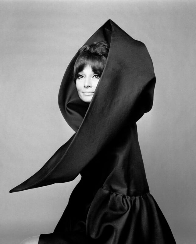 A woman poses in a dramatic black dress with an oversized, sculptural collar framing her face against a plain background, creating a striking and elegant fashion portrait.