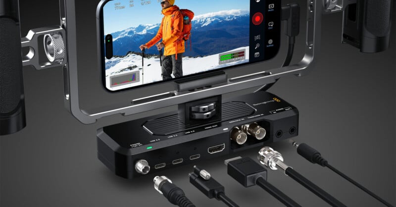 A smartphone mounted in a filming rig displays a person in an orange jacket on a snowy mountain. Below, various video and audio cables are connected to a compact black device with multiple ports and controls.