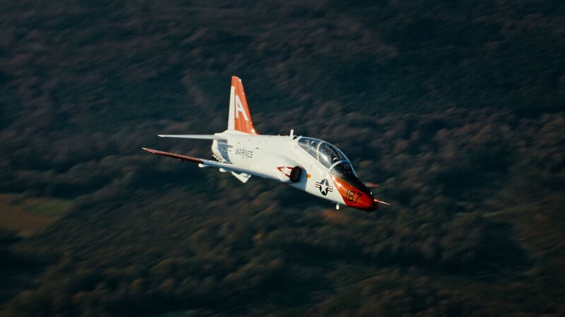 A white and red military jet aircraft with a large "A" on its tail is flying over a forested landscape.