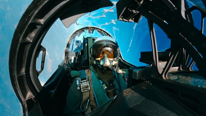 A fighter pilot wearing a helmet and oxygen mask sits in the cockpit of a jet, flying against a bright blue sky, with cockpit controls and the canopy visible around them.