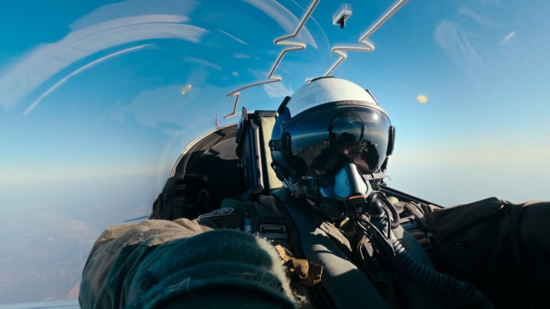 A fighter pilot wearing a helmet and oxygen mask takes a selfie inside a jet cockpit mid-flight, with a clear blue sky and cockpit reflections visible in the background.