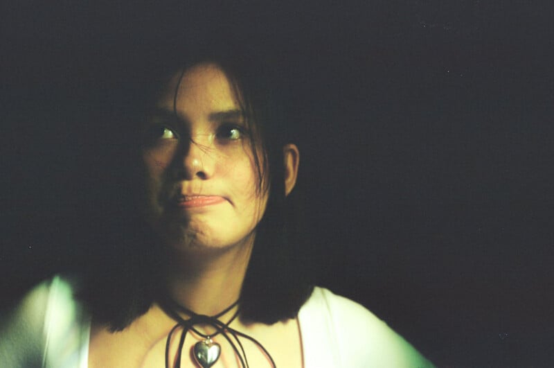 A young woman with shoulder-length dark hair looks upward with a thoughtful expression, her lips pressed together. She wears a necklace with a heart-shaped pendant and is softly lit against a dark background.