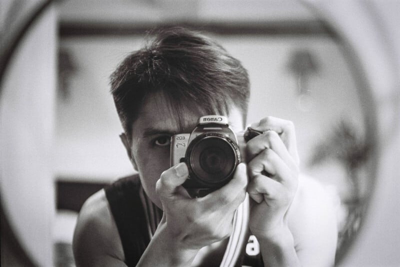 A person with short hair is taking a black-and-white photo of themselves in a mirror, holding a Canon camera close to their face. Their reflection is centered and framed by the circular mirror.
