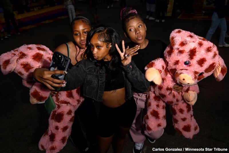 Three young women take a selfie together at night, each holding a large pink stuffed animal that resembles a giraffe. They make playful faces as they pose for the photo.
