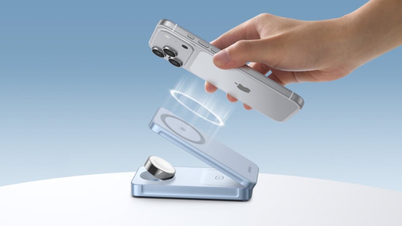 A hand holds a silver smartphone above a sleek blue wireless charging stand, with glowing rings indicating the phone is about to connect for charging. The background is a gradient of light blue.
