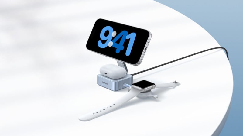 A smartphone, wireless earbuds, and a smartwatch are being charged on a sleek, compact charging station placed on a white surface. The phone displays the time 9:41.