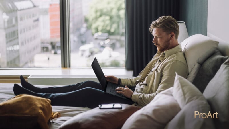 A man sits on a bed using a laptop in a bright room with large windows, city buildings and trees visible outside. A phone and orange bag are on the bed. The image is branded with the "ProArt" logo.
