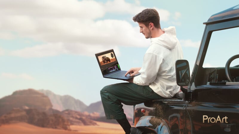 A person sits on the edge of a black vehicle in a desert landscape, using a laptop with photo editing software open. The scene includes mountains and a cloudy sky in the background. The vehicle has "ProArt" branding.