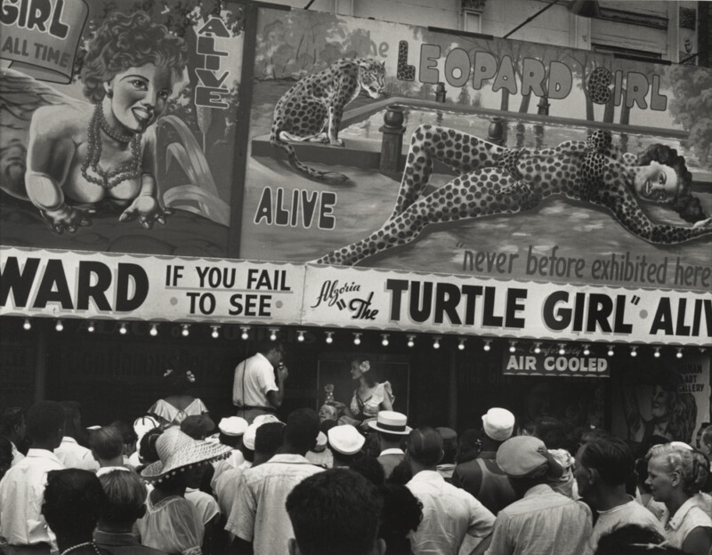 A crowd gathers in front of a carnival sideshow featuring large posters of “Leopard Girl” and “Turtle Girl,” with illustrated women in animal costumes and text advertising them as “alive” and “never before exhibited here.”.