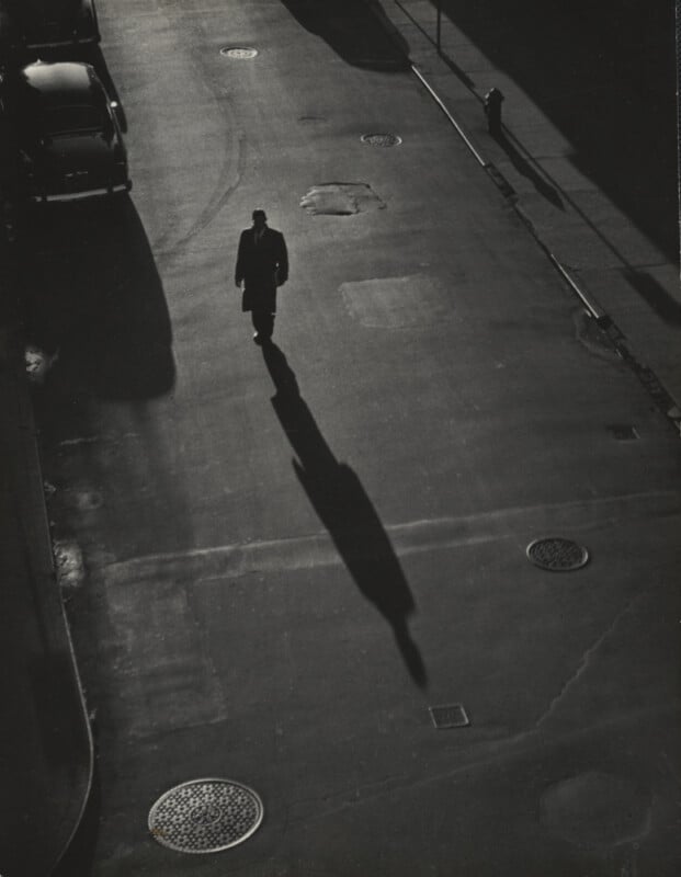 A lone person in a long coat and hat walks down an empty, shadowy street, casting an elongated shadow in the early morning or late afternoon light. A few cars, manholes, and a fire hydrant are visible.