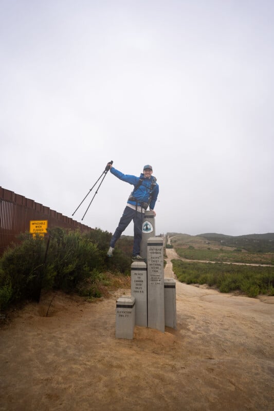 A hiker in blue gear stands on a trail marker at the start of the Pacific Crest Trail, holding trekking poles aloft. A fence and a warning sign are on the left, with a dirt path leading into the hills on a cloudy day.