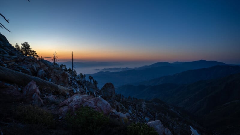 Rocky mountain landscape at sunset, with a clear sky fading from deep blue to orange, distant mountain ranges, scattered trees, and mist settling in the valleys.