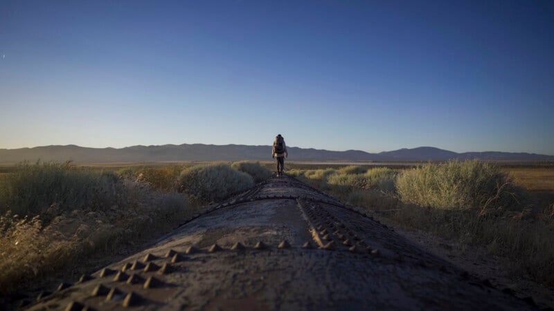 A person with a backpack walks alone on a weathered pipeline through a vast, open landscape with scrubby bushes, under a clear blue sky and distant mountains.