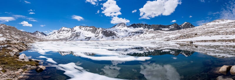 A panoramic view of a partially frozen alpine lake with snow-covered mountains in the background, reflected in the calm blue water under a bright sky with scattered clouds.