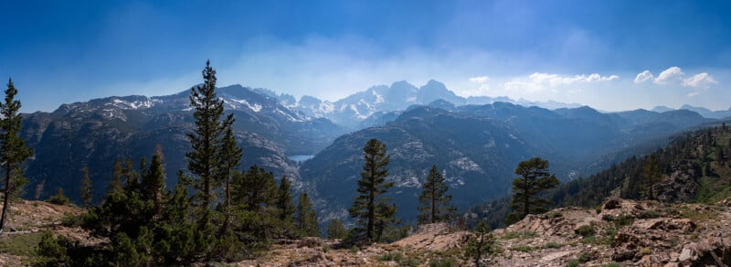 Panoramic view of a mountain range with snow-capped peaks, pine trees in the foreground, rocky terrain, and a distant lake under a clear blue sky.
