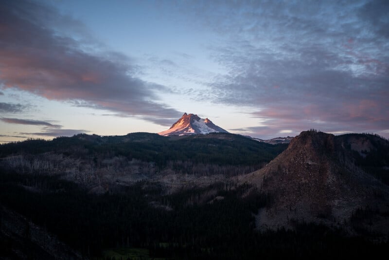 Snow-capped mountain peak illuminated by early morning or late evening sunlight, surrounded by dark, forested hills and dramatic, partly cloudy sky with pink and blue tones.