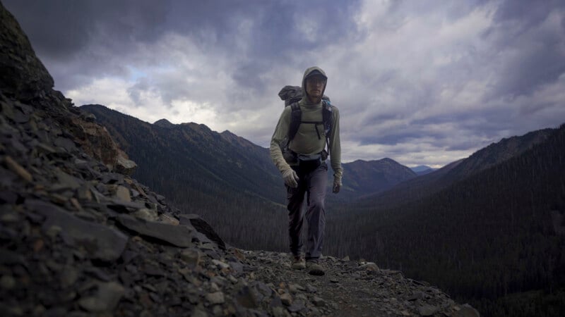 A person wearing a hooded jacket and backpack hikes up a rocky mountain trail under a cloudy sky, with forested mountains stretching into the distance.
