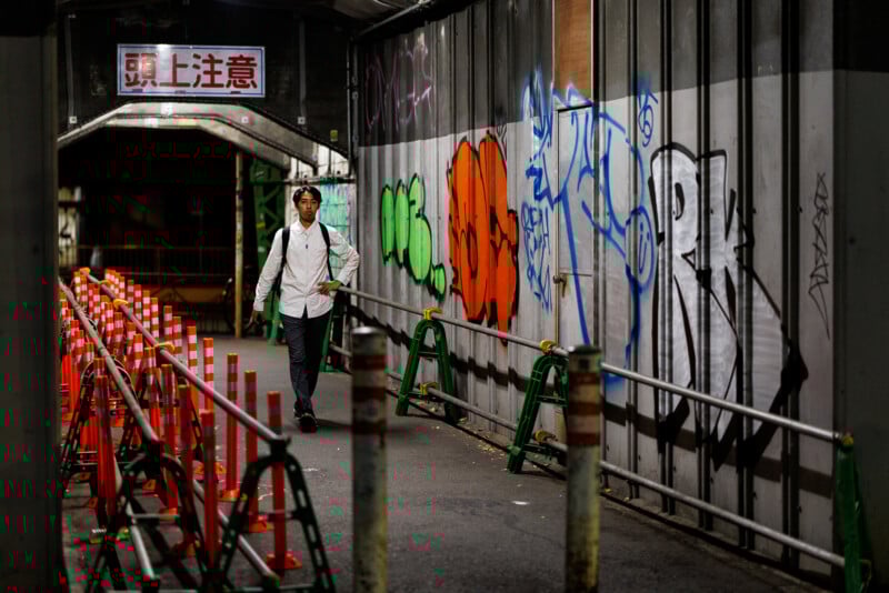 A man in a white shirt and backpack walks through a dimly lit urban passageway lined with orange traffic cones and graffiti-covered walls at night. A sign with Japanese characters hangs overhead.