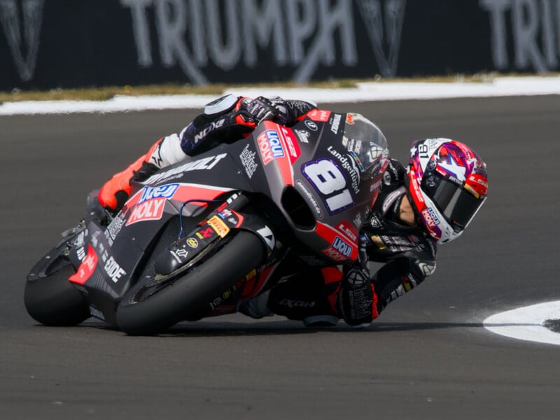 A motorcycle racer wearing a black and red suit and helmet leans sharply into a turn on a race track, riding a black bike labeled "81." The blurred Triumph logo is visible in the background.