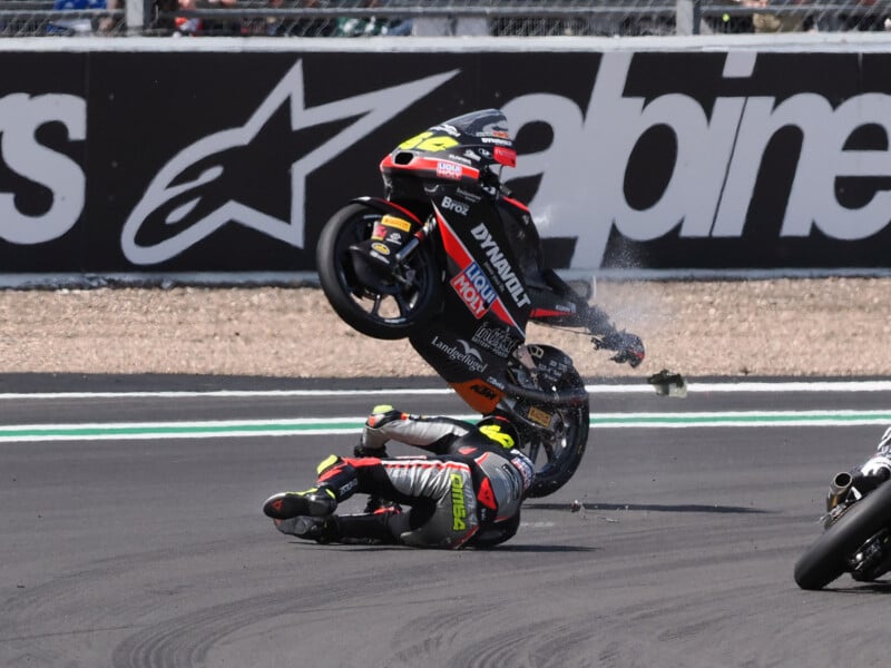 A motorcycle is airborne above a racer who has fallen on the track, with debris flying around, during a motorsport event. Safety gear and brand logos are visible on the suit and bike.