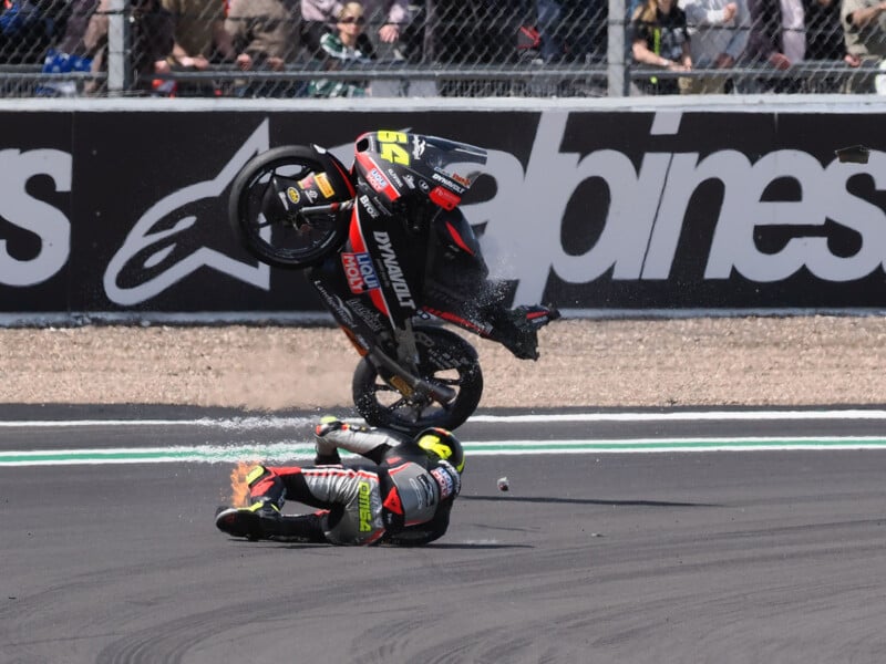 A motorcycle racer falls and slides on the track as his bike flips through the air, with debris scattering; spectators watch behind a fence in the background.