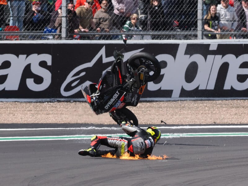 A motorcycle racer crashes on the track, sliding on the ground as their bike flips in the air above them. Flames and sparks are visible near the rider, with spectators watching from behind a fence.
