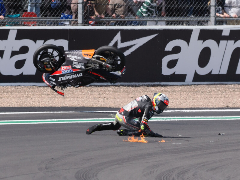 A motorcycle racer crashes on a track, sliding on the ground as his bike flips in the air behind him. A small patch of flames appears near the rider, and spectators watch from behind a fence.