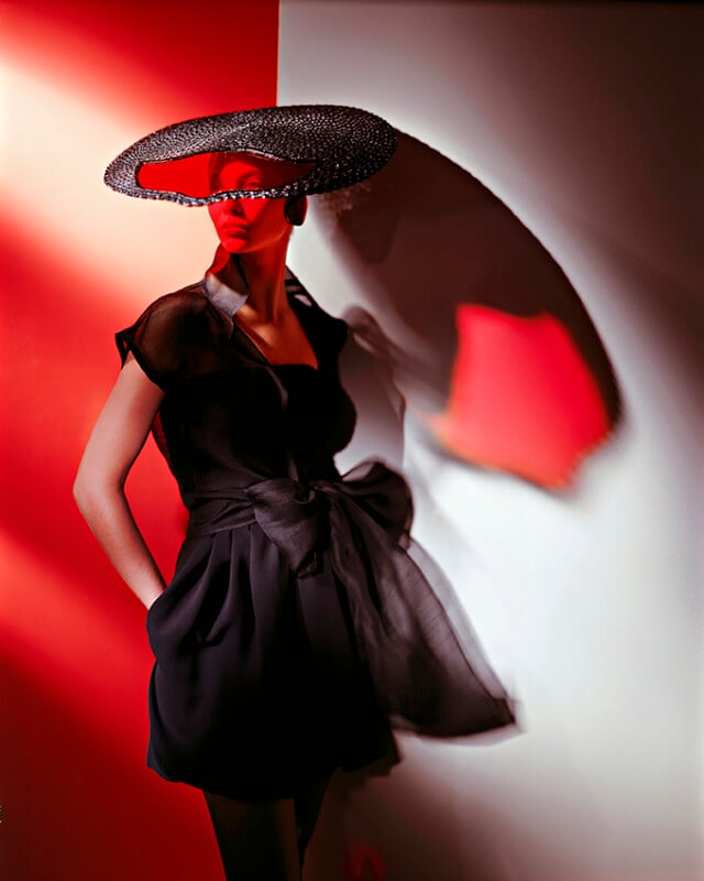 A woman in a black dress with a large bow poses against a red and white wall, wearing a wide, circular hat that casts dramatic shadows on her face and background. The lighting creates a bold, artistic effect.