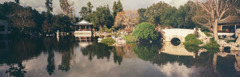 Una escena de jardín serena con un estanque con reflejos, puentes de piedra blanca, exuberantes árboles y un cenador tradicional rodeado por una porción de cielo nublado.