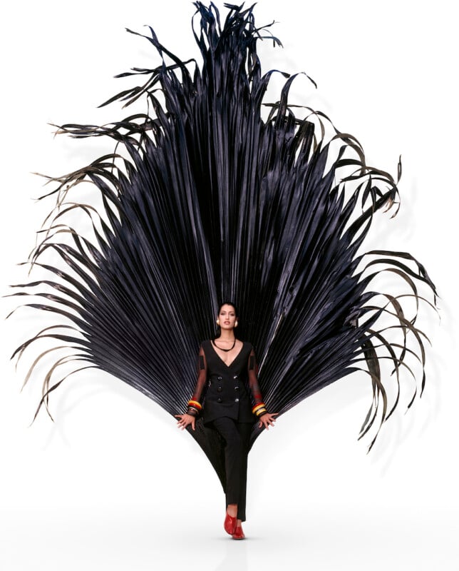 A woman in a black suit poses in front of a large, dramatic black palm frond display, which fans out behind her like wings, against a white background. She wears red shoes and colorful bangles.