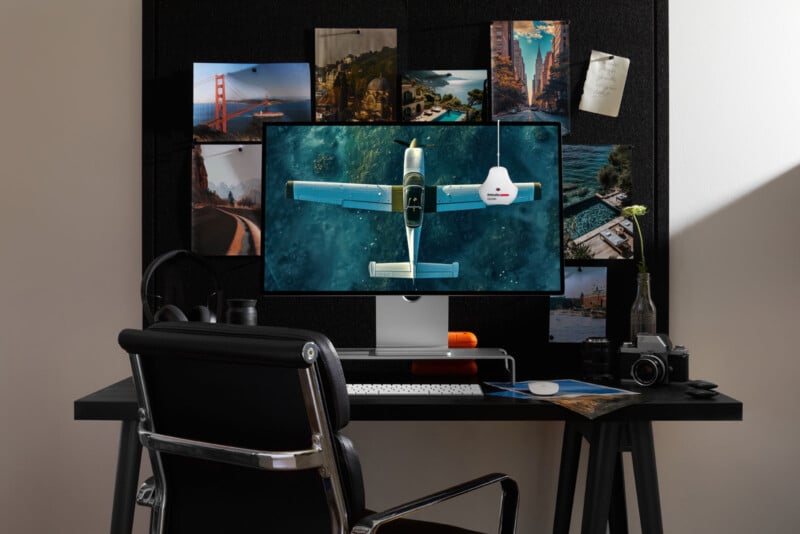 A modern desk setup with a large monitor displaying a flight simulator, surrounded by travel photos pinned on a board. The desk also holds headphones, a keyboard, a camera, and a small vase with a single flower.