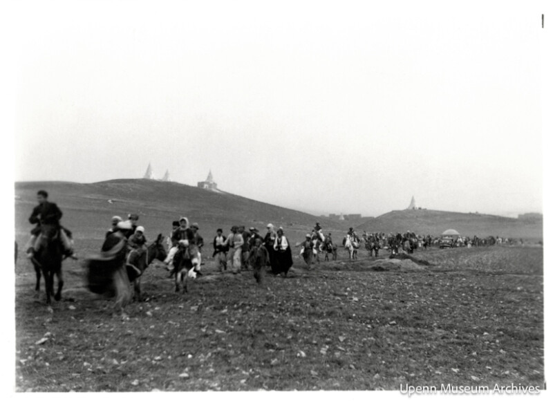 A long line of people, some on horseback and others on foot, travels across a barren, uneven landscape with hills and distant buildings in the background. The scene appears historical. "Upenn Museum Archives" is noted in the corner.
