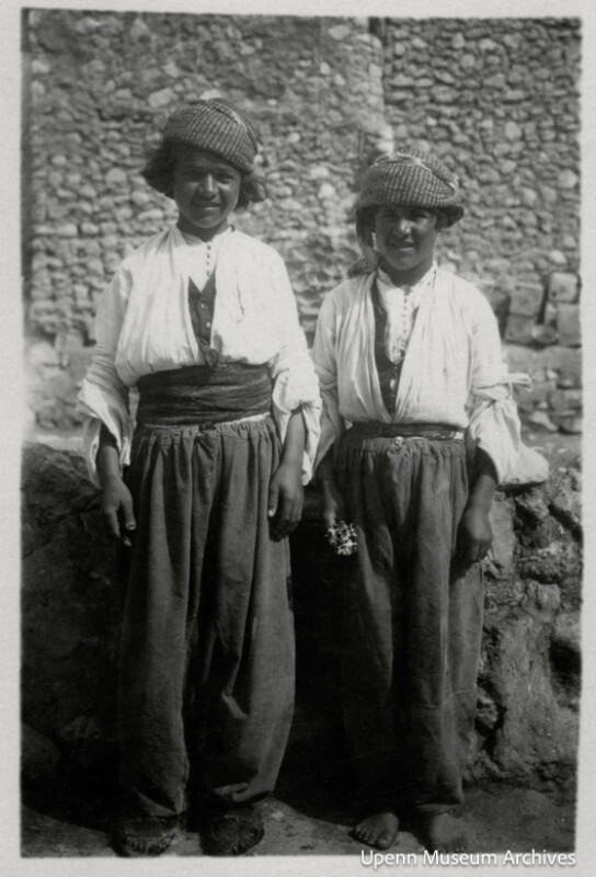 Two young people wearing traditional loose pants, belted tunics, and patterned headscarves stand side by side in front of a stone wall. Both are smiling at the camera. The image is black and white.
