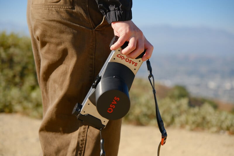 Hombre con pantalones marrones de pie al aire libre sosteniendo una cámara grande marcada "Es" En las letras rojas, hay un hermoso paisaje en el fondo.