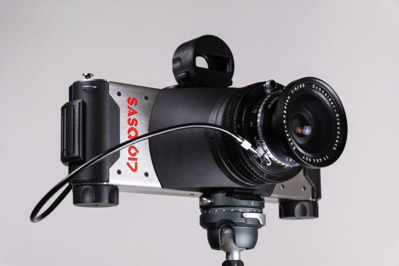 Primer plano de la cámara de película panorámica Sasqid montada en un trípode con una lente grande y una liberación de cables, ubicada sobre un fondo de gris claro puro.