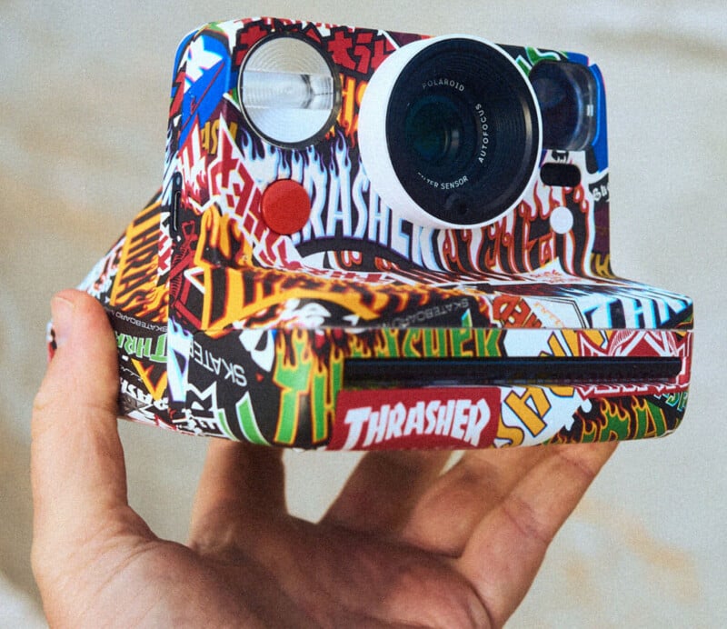 A hand holds a Polaroid camera covered in colorful, overlapping skateboard and "Thrasher" stickers, creating a bold, vibrant look. The background is softly blurred.