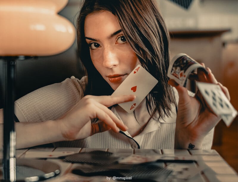 A young woman with brown hair holds playing cards, focusing intently while showing the ace of hearts. She sits indoors, with a lamp and a blurred background visible behind her.