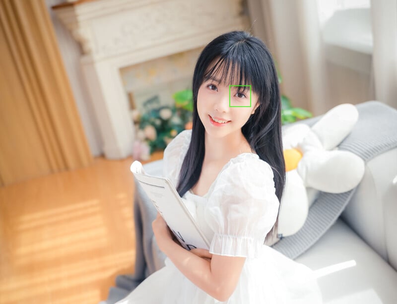 A young woman in a white dress sits on a couch, smiling and holding a clipboard. Sunlight streams through a window, and a plush toy is beside her. The room appears bright and cozy with soft, warm colors.