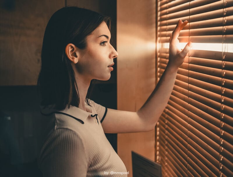 A young woman stands indoors, gazing thoughtfully through window blinds as sunlight filters in, illuminating her face and hand. She wears a short-sleeved shirt and appears calm and contemplative.