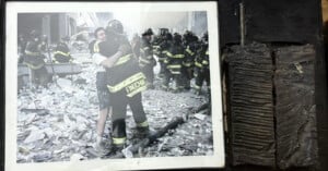 A framed photo shows two men—one in firefighting gear—embracing amid debris, while several firefighters stand behind them. To the right of the photo is a charred, ribbed metal object.