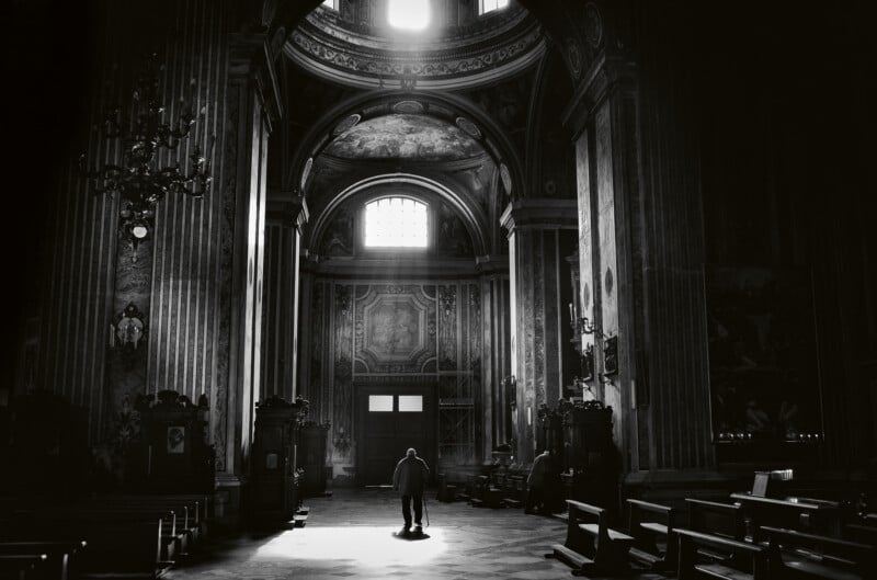 A person stands alone in the center of a grand, dimly lit cathedral with high arches, columns, ornate decorations, and sunlight streaming through a high window.