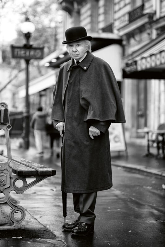 An older person in a long cloak and bowler hat stands on a wet city sidewalk, holding a cane. Parisian architecture and a "Metro" sign appear in the background. The image is in black and white.