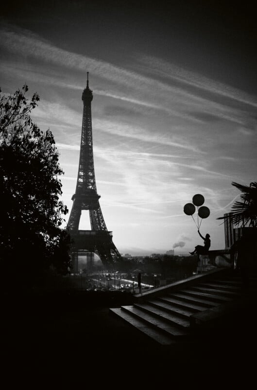 A black and white photo of the Eiffel Tower at dusk with a silhouetted person holding balloons, sitting on a stair railing in the foreground. Trees frame the scene, creating a dreamy, serene Parisian atmosphere.
