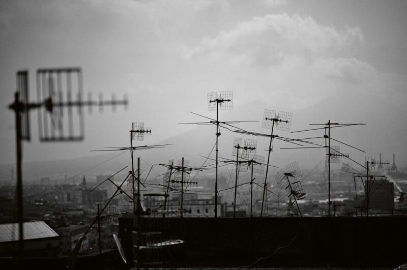 A cluster of TV antennas on rooftops overlook a hazy cityscape, with distant buildings barely visible through the mist under a cloudy sky. The image is in black and white.