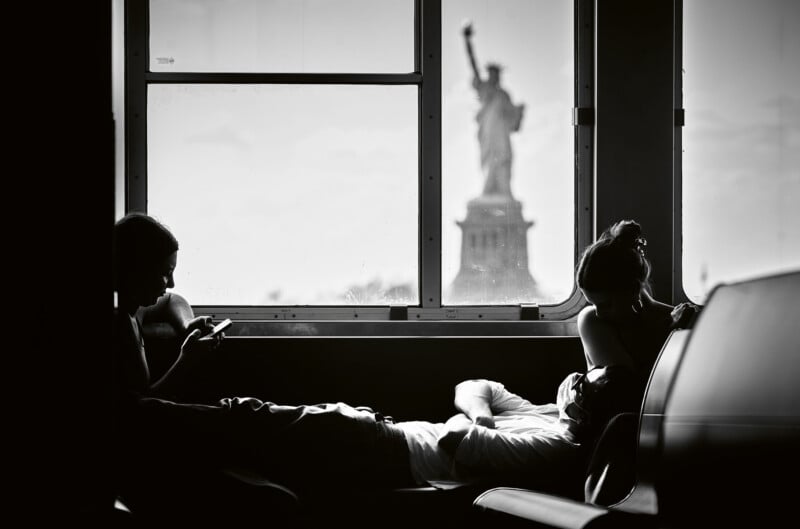 Three people relax on a bench by a window; one uses a phone, another reclines with eyes closed, and the third looks down. The Statue of Liberty is visible through the window in the background.