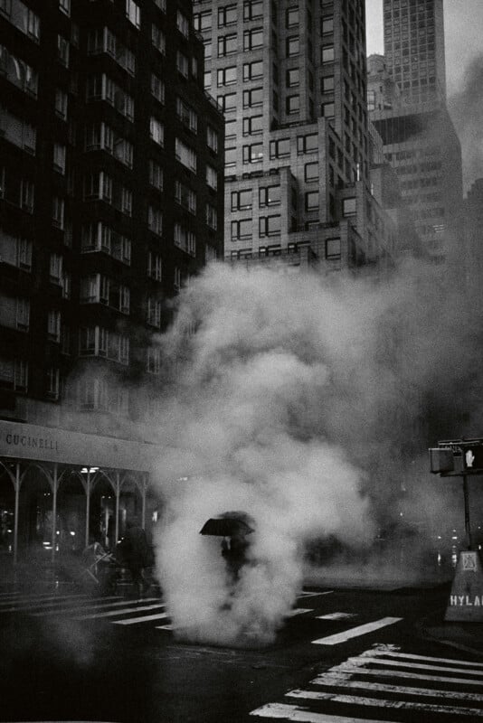 A person holding an umbrella crosses a city street enveloped in thick steam, surrounded by tall buildings in a moody, black-and-white urban scene.