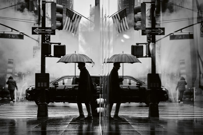A person holding an umbrella crosses a city street on a rainy day, their silhouette and surroundings reflected in a glass wall. Steam rises from the ground, and American flags and street signs are visible.