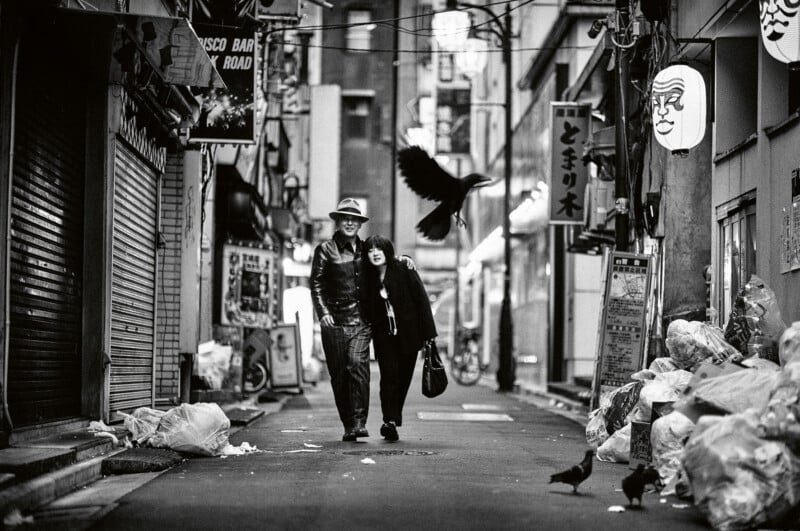 A couple walks arm in arm down a narrow alley lined with garbage bags and closed shop fronts, as crows fly and scavenge nearby; Japanese signs hang overhead. The scene is in black and white.
