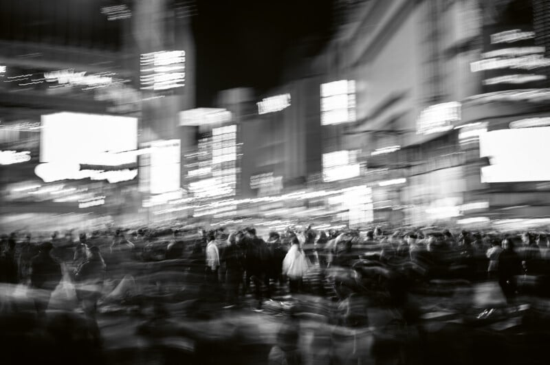 A black and white photo shows a busy city street at night, crowded with people. The image is intentionally blurred, creating a sense of motion and energy with streaks of lights and indistinct figures.